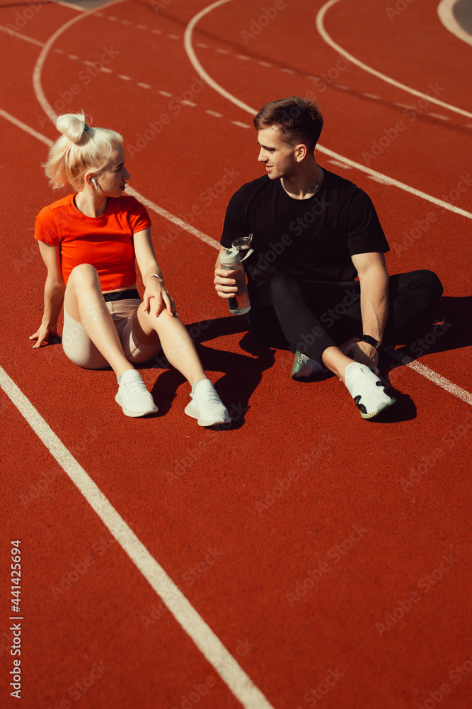 young beautiful blonde and a guy are sitting on a jogging track with a bottle of water and chatting young beautiful blonde and a guy are sitting on a jogging track with a bottle of water and chatting