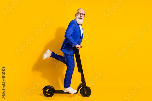 Full body profile photo of funny grey beard aged man ride scooter wear spectacles blue suit isolated on yellow background photo