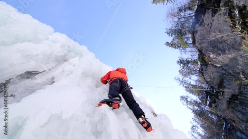 Low angle shot of a male athlete climb cliff covered with ice, swinging the ice axe and using crampons to get a foothold