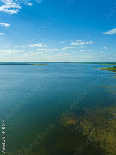 View from the drone is of a blue lake with a reflection of the sun and a green forest in the distance. Beautiful landscape from a drone on a sunny day. Vrtical photo