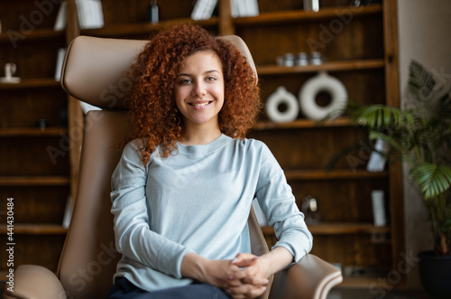 Attractive red-haired woman sitting on the armchair and looks at the camera, portrait of beautiful girl with toothy smile relaxing in the armchair at home or office