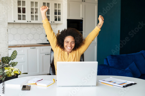 Overjoyed African-American freelancer woman yelling yes, looking at the laptop screen, celebrating victory, goal achievement, black female with afro hairstyle raised arms up, received opportunity photo