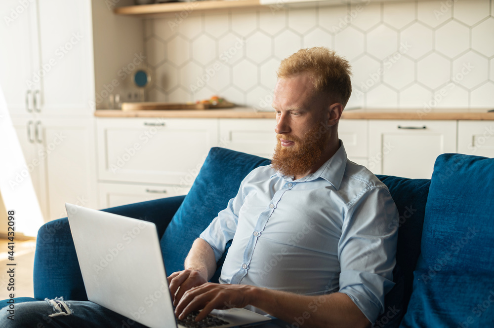 Focused redhead man using laptop computer for studying or working online, freelance guy typing sitting on the sofa at home, develops new project working remotely Focused redhead man using laptop computer for studying or working online, freelance guy typing sitting on the sofa at home, develops new project working remotely