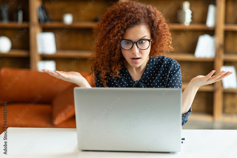 Puzzled and confused redhead female entrepreneur staring at the laptop screen, curly woman in shirt and eyewear looking at monitor with asking expression, misunderstanding what happened