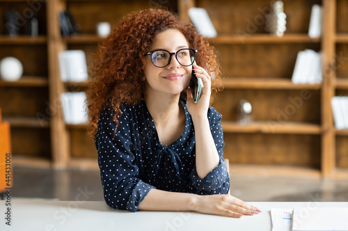 Beautiful female office employee communication on mobile phone sitting at the desk, young curly red-haired woman talking on the smartphone. People and communication concept