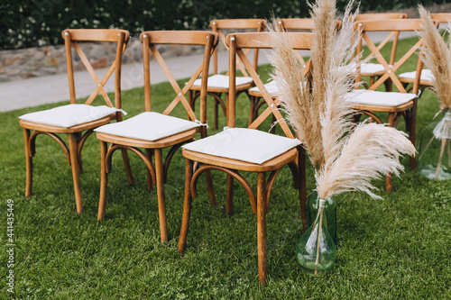 A row of wooden chairs with reed decorations stands on the green grass in the garden. Wedding photography.