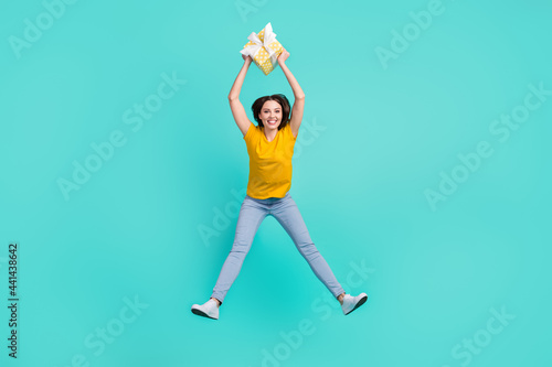 Full size photo of cool young lady jump hold present wear yellow t-shirt jeans isolated on teal color background