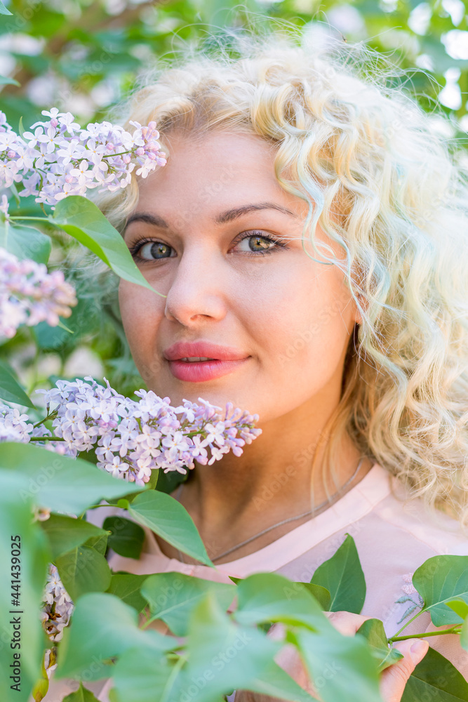 Obraz premium A beautiful young and happy blonde girl stands next to a blooming lilac tree. Relaxation in the spring garden.