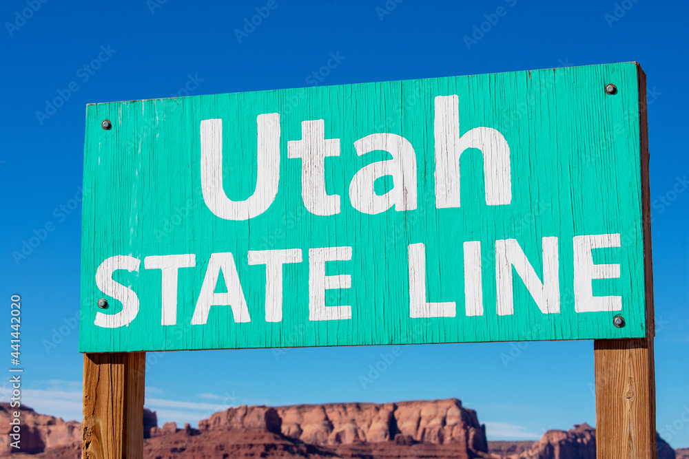 Utah state line weathered plywood sign on wood posts. Blurred scenic dry desert landscape with butte mountains under blue sky Utah state line weathered plywood sign on wood posts. Blurred scenic dry desert landscape with butte mountains under blue sky