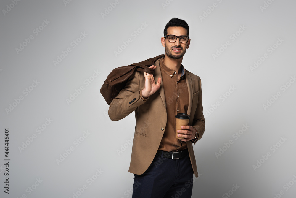 Smiling arabian businessman holding coffee to go and handbag isolated on grey Smiling arabian businessman holding coffee to go and handbag isolated on grey
