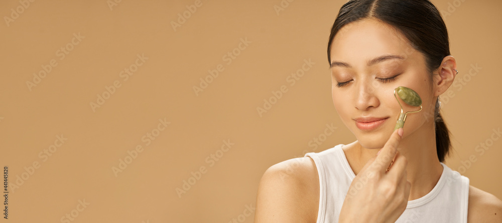 Obraz na płótnie Website header of Facial beauty treatment. Beautiful young woman looking relaxed while getting massage face using jade roller isolated over beige background w salonie
