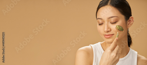Website header of Facial beauty treatment. Beautiful young woman looking relaxed while getting massage face using jade roller isolated over beige background