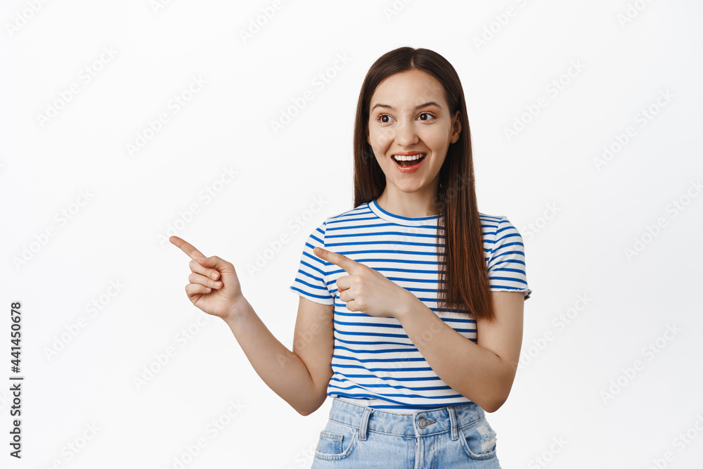 Happy girl client pointing fingers and looking left at sale banner, showing advertisement, laughing and smiling at event logo, standing against white background Happy girl client pointing fingers and looking left at sale banner, showing advertisement, laughing and smiling at event logo, standing against white background