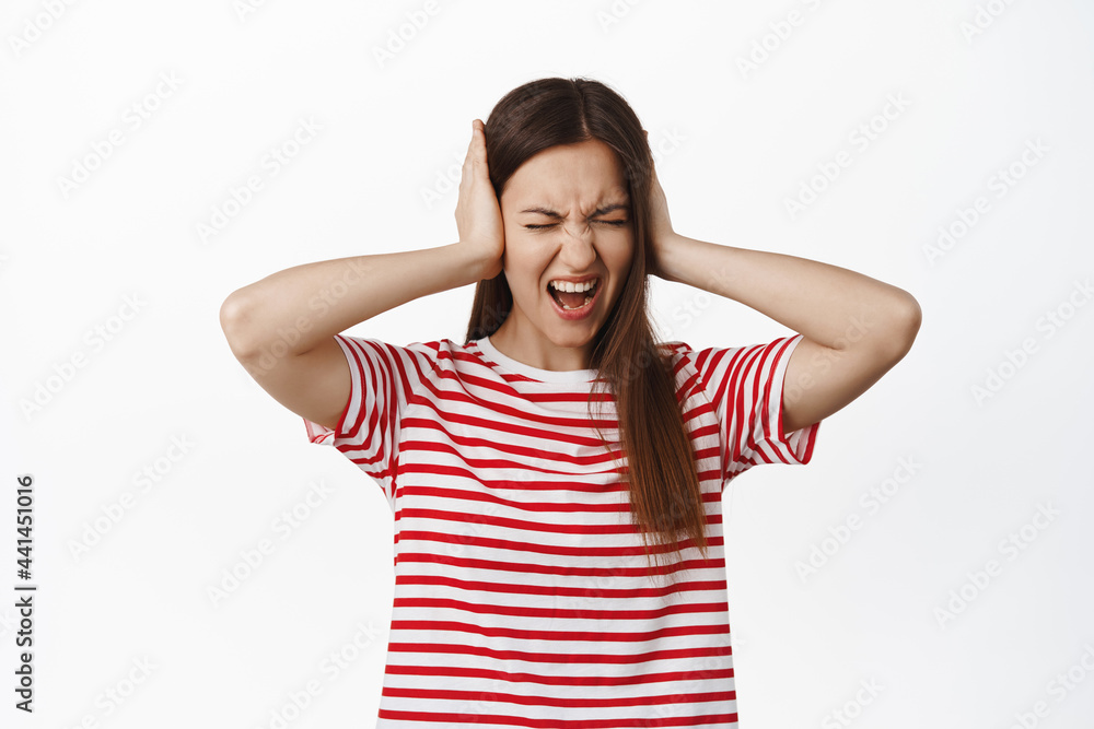 Frustrated and annoyed young woman screaming in denial, unwilling to listen, fed up with annoying loud noise, cover ears with hands and shouting, standing over white background Frustrated and annoyed young woman screaming in denial, unwilling to listen, fed up with annoying loud noise, cover ears with hands and shouting, standing over white background