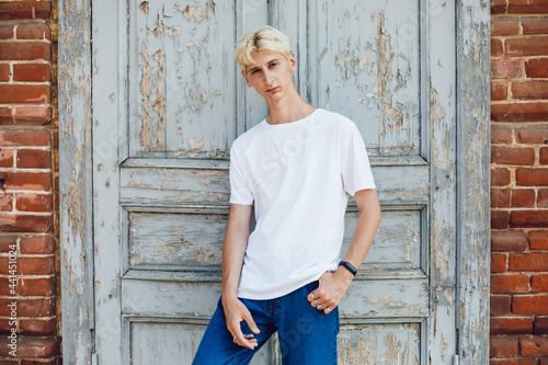 A young blond man in a white t-shirt stands against the background of an old wooden door.