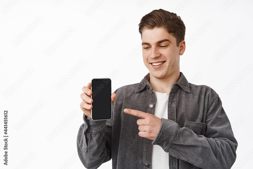 Close up of cheerful caucasian guy pointing finger at smartphone, showing mobile phone screen app, interface application, standing against white background Close up of cheerful caucasian guy pointing finger at smartphone, showing mobile phone screen app, interface application, standing against white background