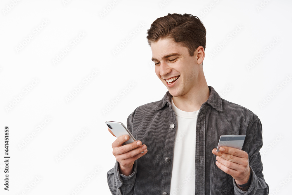 Portrait of smiling young man purchasing in mobile phone store, paying with smartphone and credit card, order delivery on cellphone, standing against white background Portrait of smiling young man purchasing in mobile phone store, paying with smartphone and credit card, order delivery on cellphone, standing against white background
