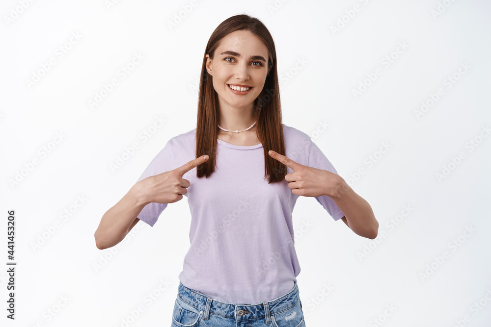 Portrait of young smiling woman, recruit pointing at herself and looking confident, choose pick me, self promoting, volunteering, standing against white background Portrait of young smiling woman, recruit pointing at herself and looking confident, choose pick me, self promoting, volunteering, standing against white background