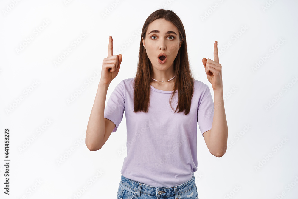 Portrait of surprised girl pointing fingers up, gasping and looking worried, showing announcement, asks question about promo offer above, white background Portrait of surprised girl pointing fingers up, gasping and looking worried, showing announcement, asks question about promo offer above, white background
