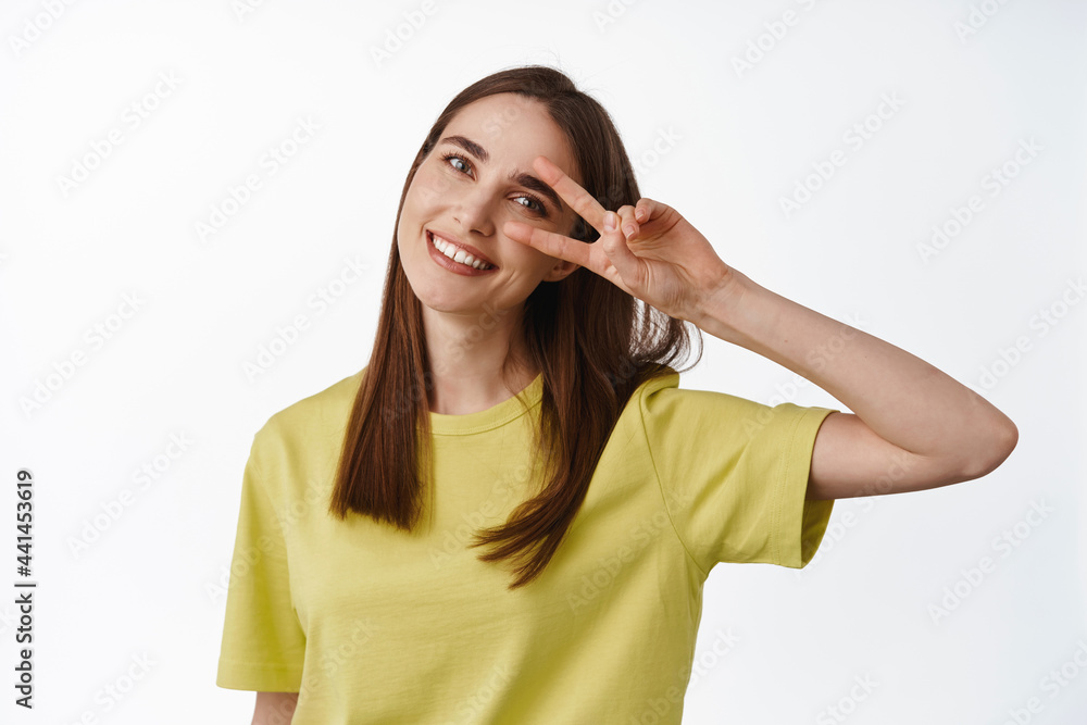 Close up portrait of beautiful girl shows peace v-sign gesture near eye, tilt head and smiling, keeping positive attitude, enjoy summer, standing over white background Close up portrait of beautiful girl shows peace v-sign gesture near eye, tilt head and smiling, keeping positive attitude, enjoy summer, standing over white background