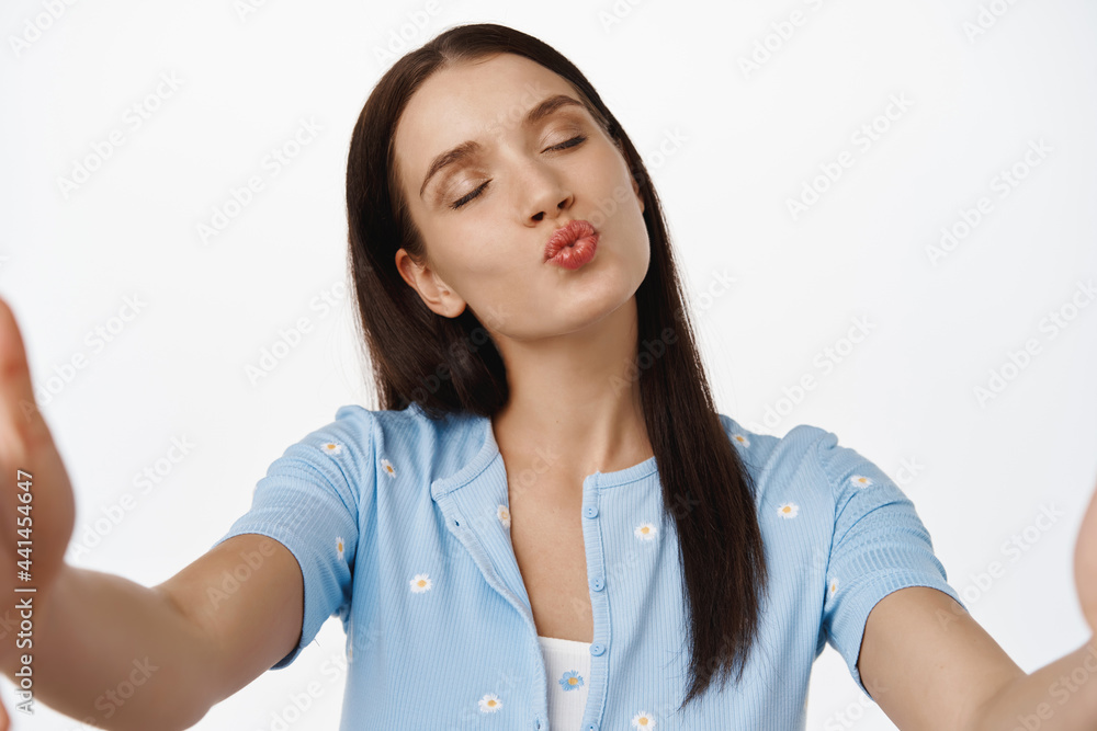 Young woman holding camera, smartphone or tablet, video calling with love partner, sending air kiss with close eyes and flirty coquettish face expression, white background Young woman holding camera, smartphone or tablet, video calling with love partner, sending air kiss with close eyes and flirty coquettish face expression, white background