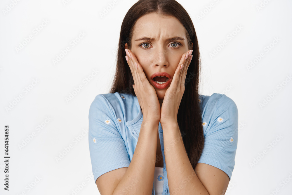 Close up of shocked and concerned woman holding hands on face, open mouth and furrow eyebrows startled, hear awful terrible news, standing against white background Close up of shocked and concerned woman holding hands on face, open mouth and furrow eyebrows startled, hear awful terrible news, standing against white background