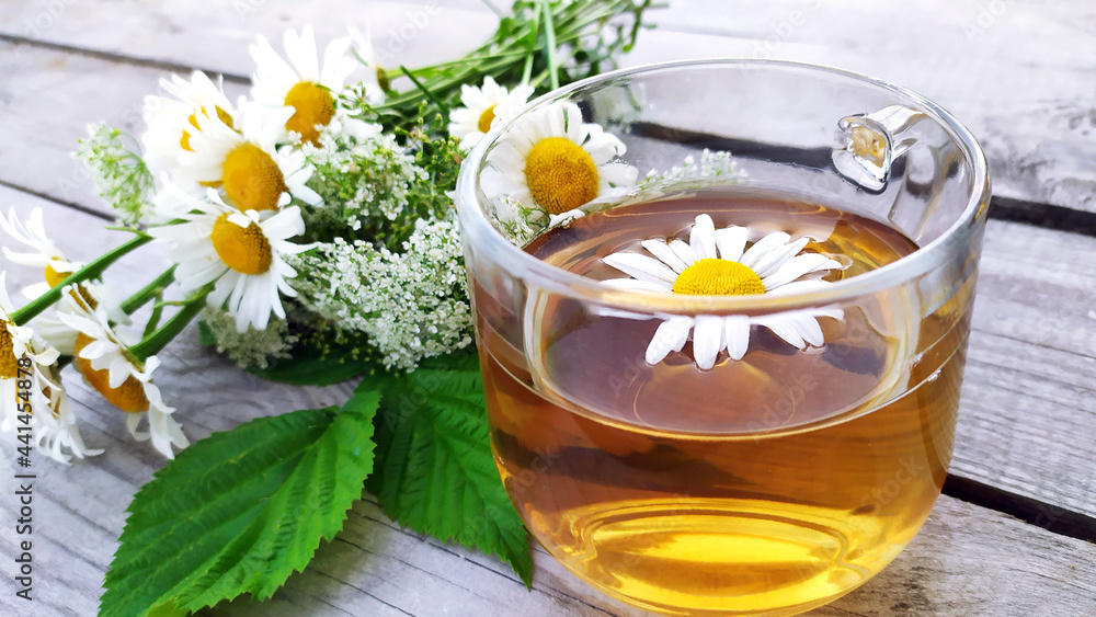 Chamomile tea in a glass cup. Floral background. A bouquet of wildflowers in the background. A transparent cup with a chamomile drink stands on a wooden background. Chamomile tea in a glass cup. Floral background. A bouquet of wildflowers in the background. A transparent cup with a chamomile drink stands on a wooden background.