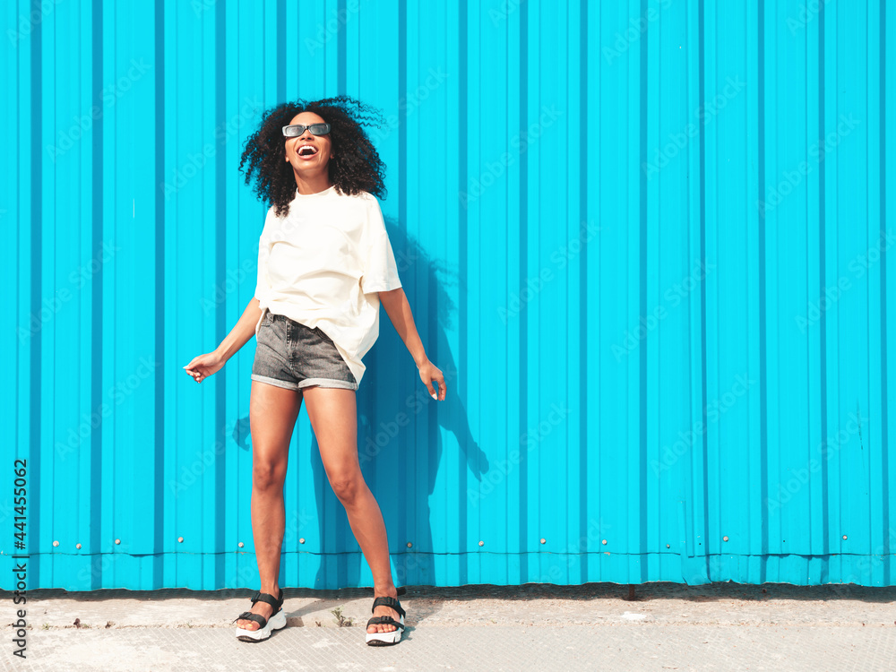 Obraz premium Beautiful black woman with afro curls hairstyle.Smiling hipster model in white t-shirt. Sexy carefree female posing in the street near blue wall in sunglasses. Cheerful and happy