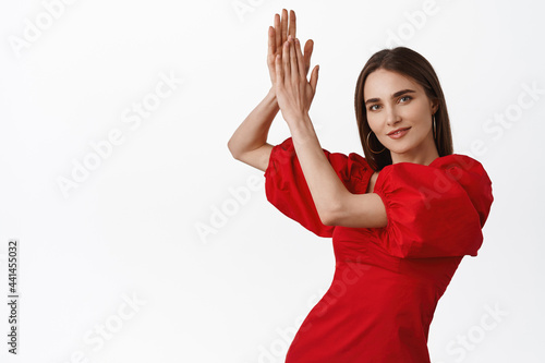 Gorgeous young hispanic woman dancing hot and sensual flamenco dance, clap in hands above head and posing in red romantic dress, smiling pleased at camera, white background