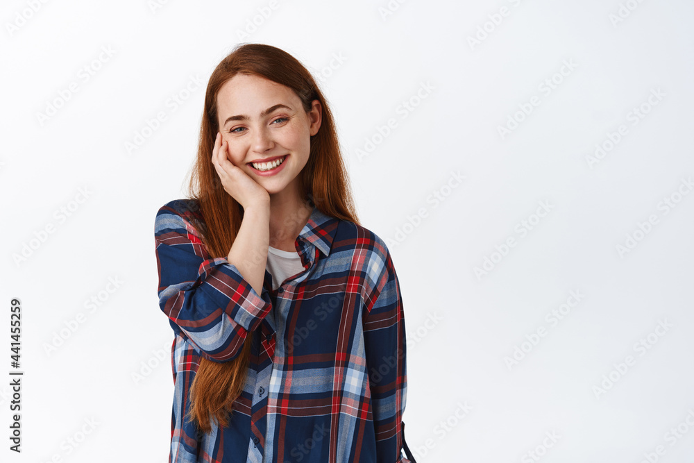 Lovely romantic girl with ginger long hair, blushing and smiling delighted, looking happy and pleased, standing against white background
