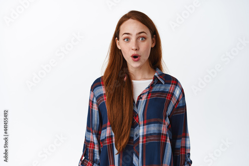 Wow super cool. Impressed teen redhead girl, gasp and stare astoished at camera, looking with excitement at awesome promo in store, standing over white background © Cookie Studio