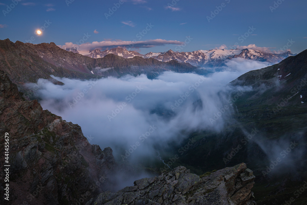 Mountain peaks emerging from the fog at the blue hour while the moon rises, Stelvio National Park, Ponte di Legno, Lombardy, Italy