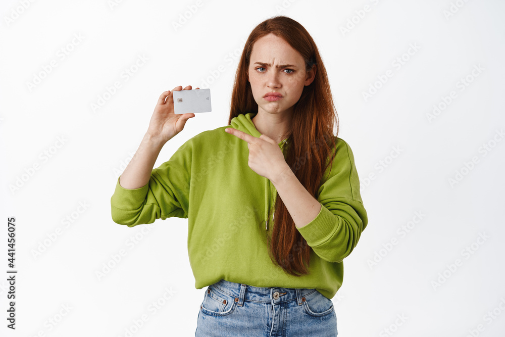 Frustrated redhead college girl pointing at credit card, frowning and looking upset, complaining, standing in casual outfit against white background Frustrated redhead college girl pointing at credit card, frowning and looking upset, complaining, standing in casual outfit against white background