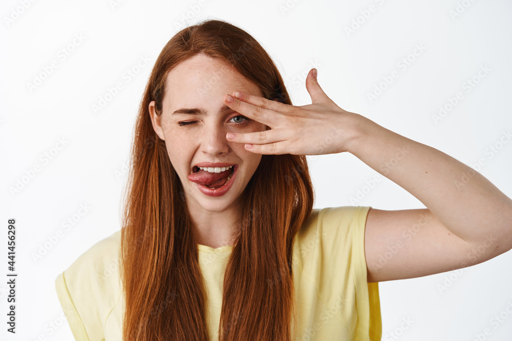 Close up portrait of happy cute girl with long red hiar, winking and showing tongue, open eye peek through fingers and smiling, standing over white background Close up portrait of happy cute girl with long red hiar, winking and showing tongue, open eye peek through fingers and smiling, standing over white background