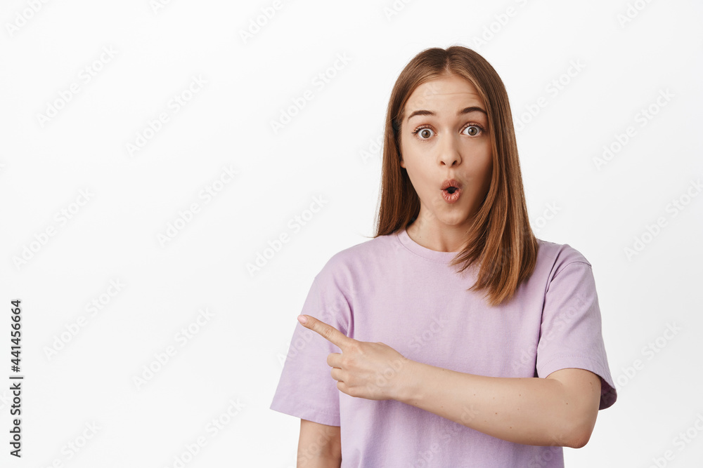 Portrait of girl looks surpirsed, says wow and points left at awesome discounts, shopping offer or logo aside copy space, standing against white background Portrait of girl looks surpirsed, says wow and points left at awesome discounts, shopping offer or logo aside copy space, standing against white background