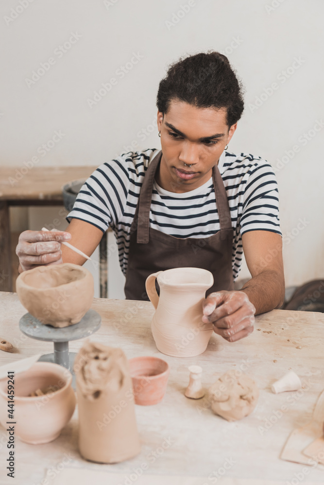 serious young african american man sculpting clay pot and stick on table with equipment in pottery