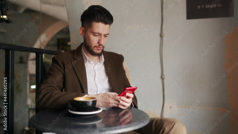 Classy young man using smartphone sitting in cafe. Guy browsing social network app in coffeshop. Office worker resting on break with a cup of coffee. Indoor scene. Classy young man using smartphone sitting in cafe. Guy browsing social network app in coffeshop. Office worker resting on break with a cup of coffee. Indoor scene.