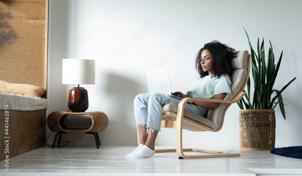 Beautiful young smiling african american woman working on laptop while sitting in bcomfortable chair at home Beautiful young smiling african american woman working on laptop while sitting in bcomfortable chair at home