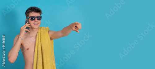 young male teenager pointing with mobile phone towel and sunglasses isolated on blue color background