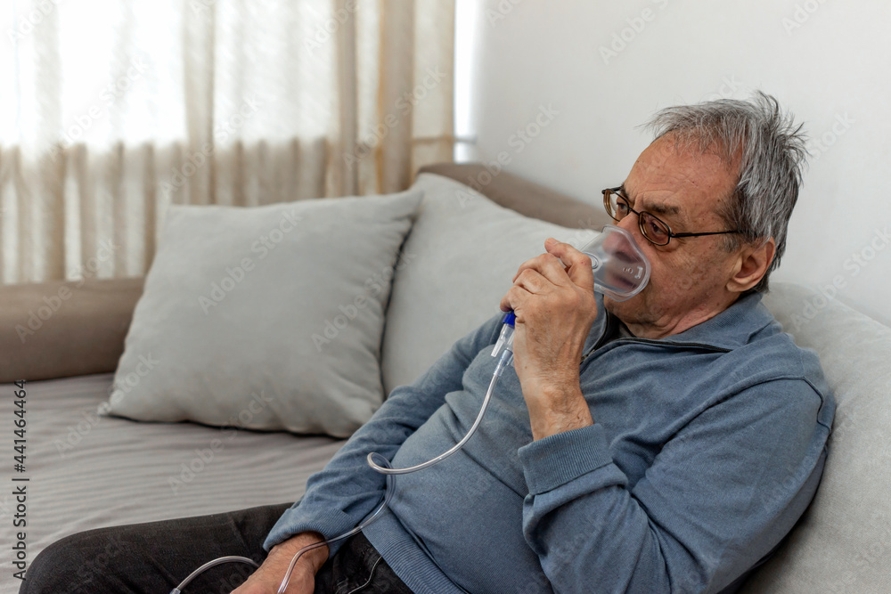 Senior man using medical equipment for inhalation with respiratory mask. Old man with serious pneumonia symptoms caused by Covid-19 virus infection sitting in home during lockdown, wearing oxygen mask Senior man using medical equipment for inhalation with respiratory mask. Old man with serious pneumonia symptoms caused by Covid-19 virus infection sitting in home during lockdown, wearing oxygen mask