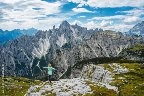 Aerial view of a woman hiker in green jacket with raised hands admiring Cadini di Misurina mountain peaks of Italian Alps, Dolomites, Italy, Europe