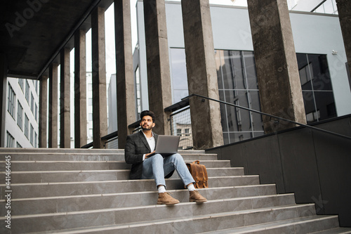 Confident hindu man in stylish clothes working on wireless laptop outdoors. Handsome young freelancer sitting on stairs and looking aside.