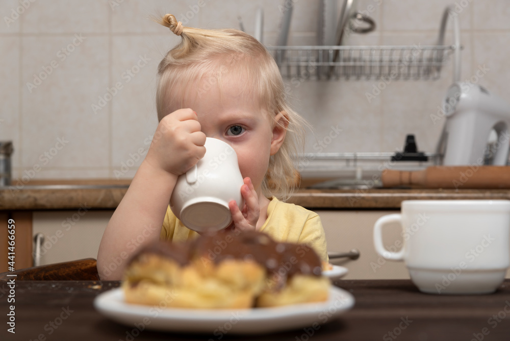 Blonde child with cup in hands in kitchen. Little smiling girl drinks a milk. Breakfast with child. Blonde child with cup in hands in kitchen. Little smiling girl drinks a milk. Breakfast with child.