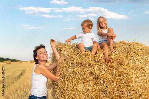 Young adult attractive beautiful mom with little son and daughter enjoy having fun fooling around sitting near golden hay bale on wheat harvested field near farm. Happy children on rural landscape