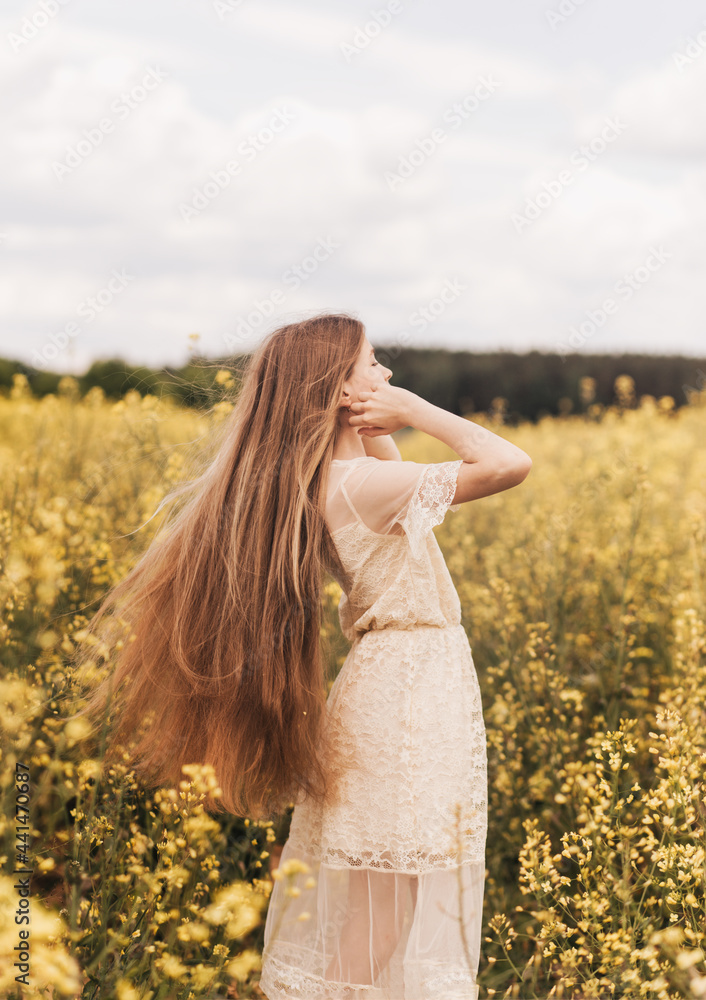 A young beautiful girl with long hair walks in a rapeseed field, looks into the distance. rear view. Vertical A young beautiful girl with long hair walks in a rapeseed field, looks into the distance. rear view. Vertical