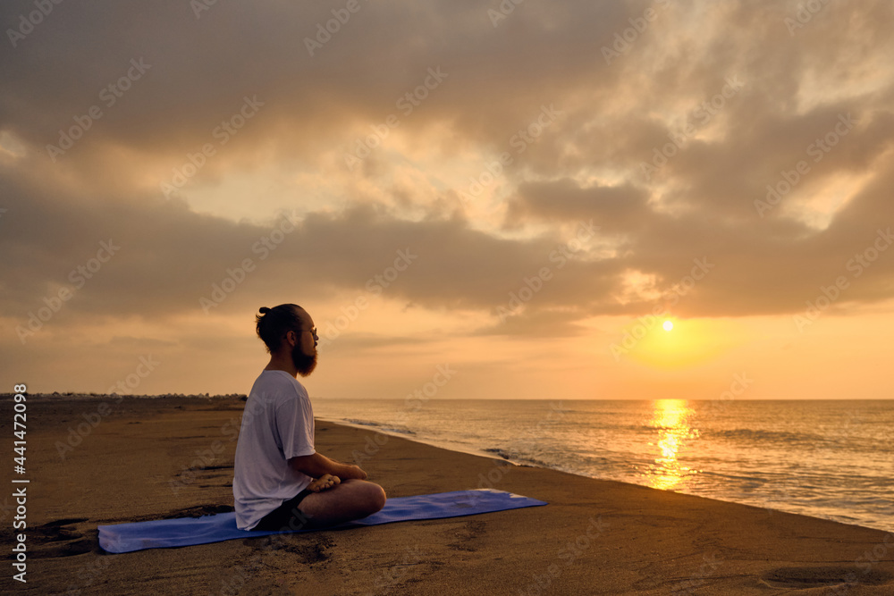 Man practices yoga meditation sitting in padmasana during sunrise. Man sits in lotus position at beach opposite rising sun and its reflection in sea Man practices yoga meditation sitting in padmasana during sunrise. Man sits in lotus position at beach opposite rising sun and its reflection in sea