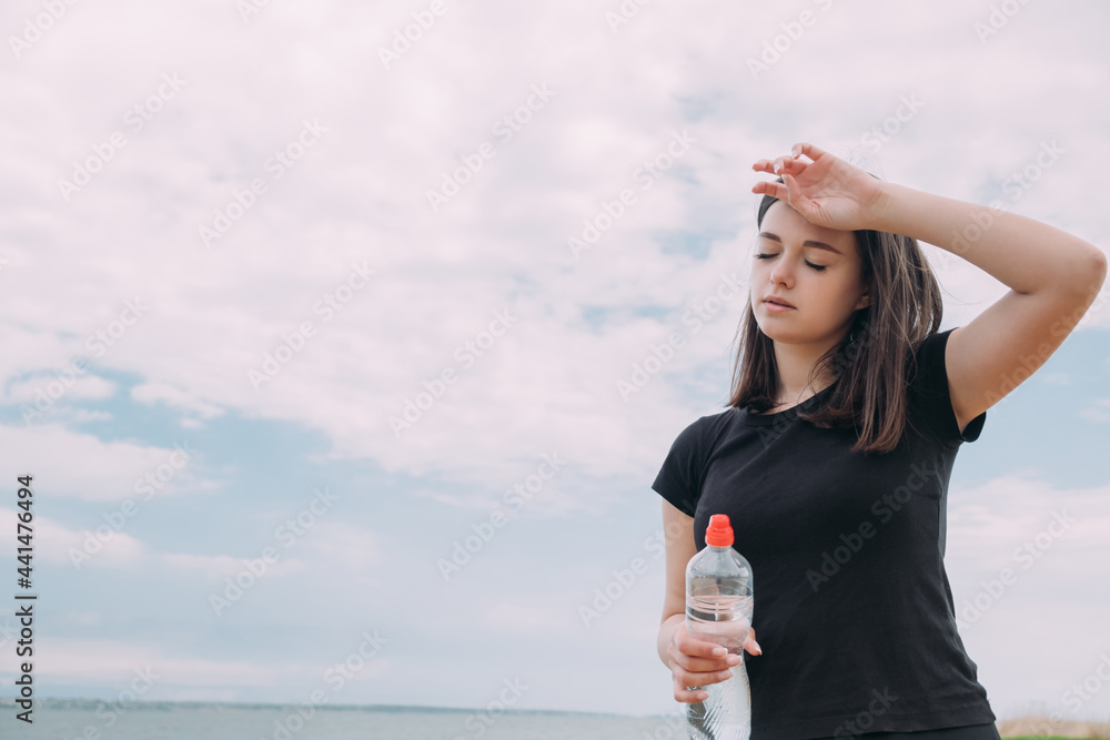 A girl of Caucasian appearance, after jogging, wipes sweat from her forehead and holds a bottle of water against the background of the sky. Outdoor sports concept A girl of Caucasian appearance, after jogging, wipes sweat from her forehead and holds a bottle of water against the background of the sky. Outdoor sports concept