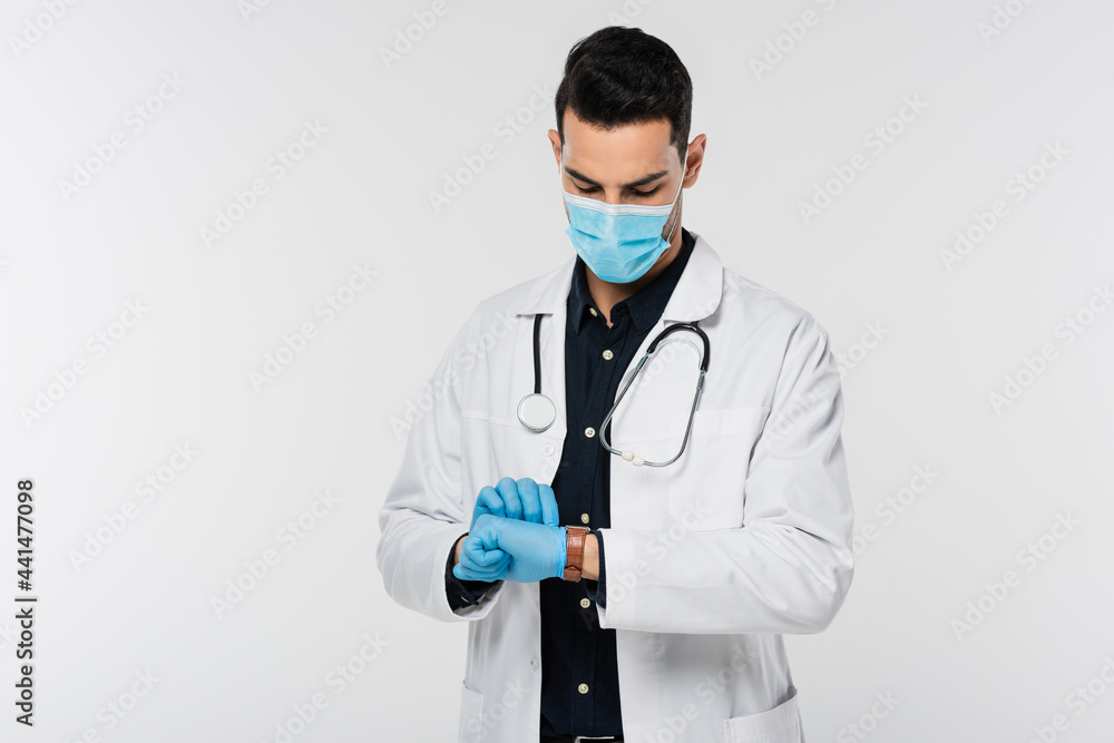 Muslim doctor in medical mask and latex gloves looking at wristwatch isolated on grey Muslim doctor in medical mask and latex gloves looking at wristwatch isolated on grey