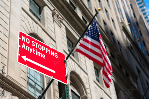No Stopping Anytime red warning road sign with an arrow. The flag of The United States Of America. Tall buildings of an urban landscape. © MichaelVi