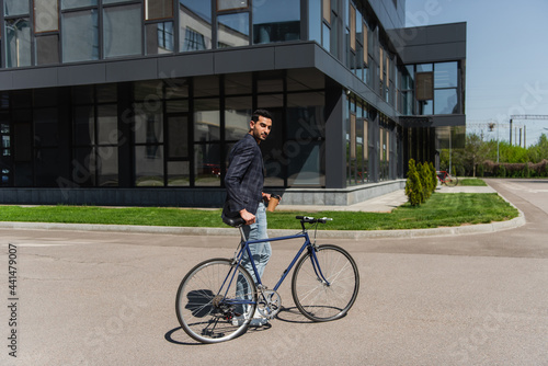 Arabian businessman holding paper cup near bike on urban street